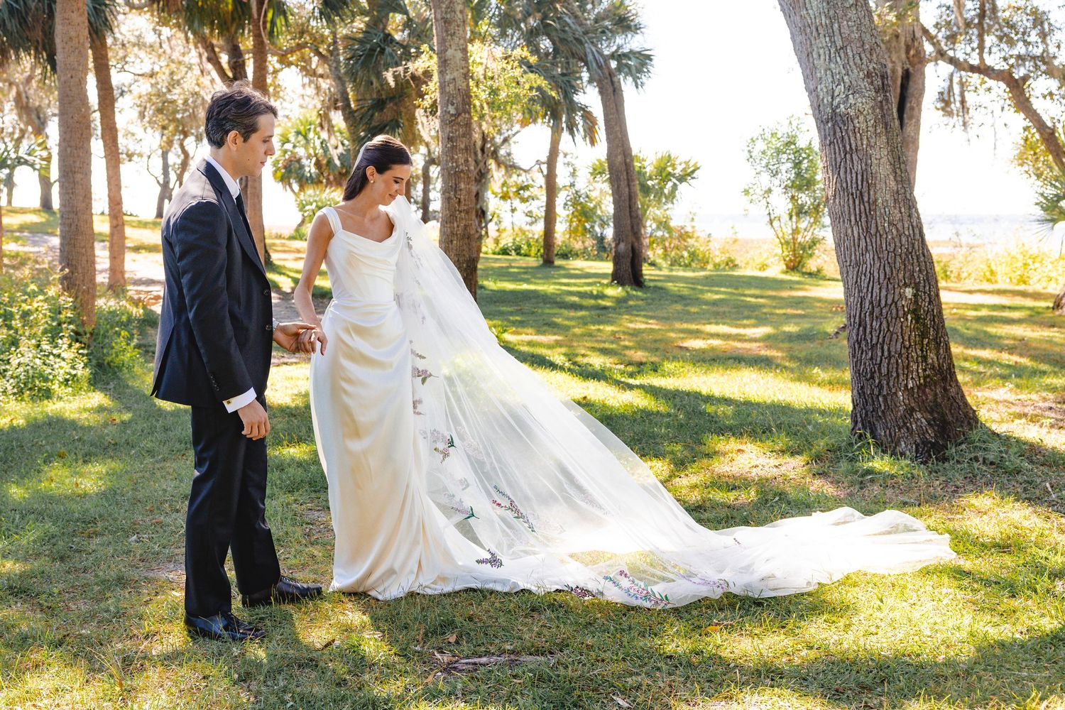 A couple in wedding attire embraces while walking through a sunlit park with tall palm trees and pine trees.