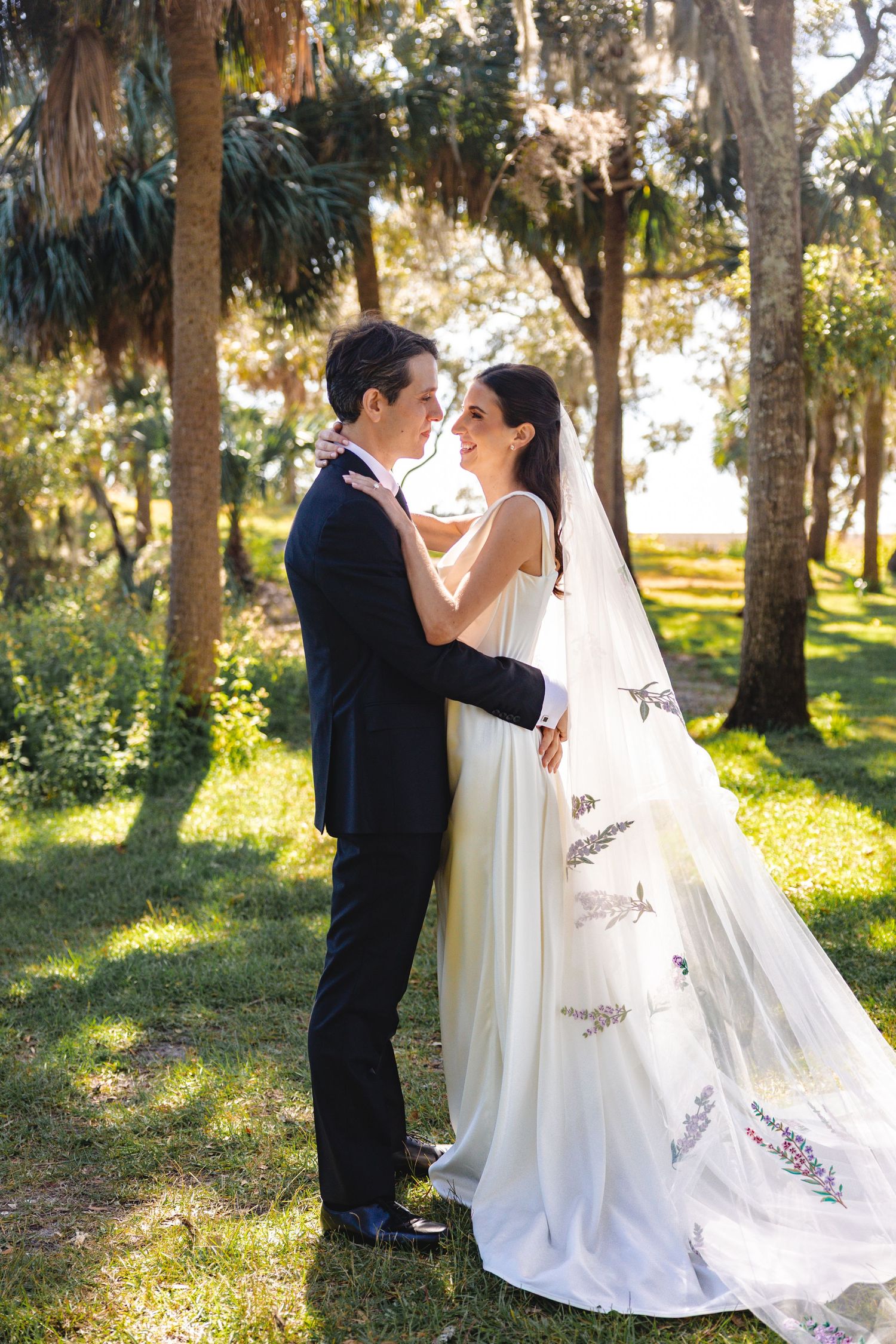 A romantic wedding couple shares intimate moments in a sunlit garden setting with tall trees.