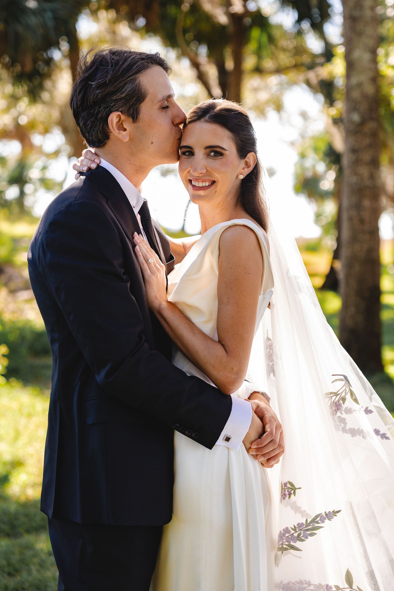 Romantic wedding couple shares intimate moments in a sunlit forest setting with flowing white dress and veil.