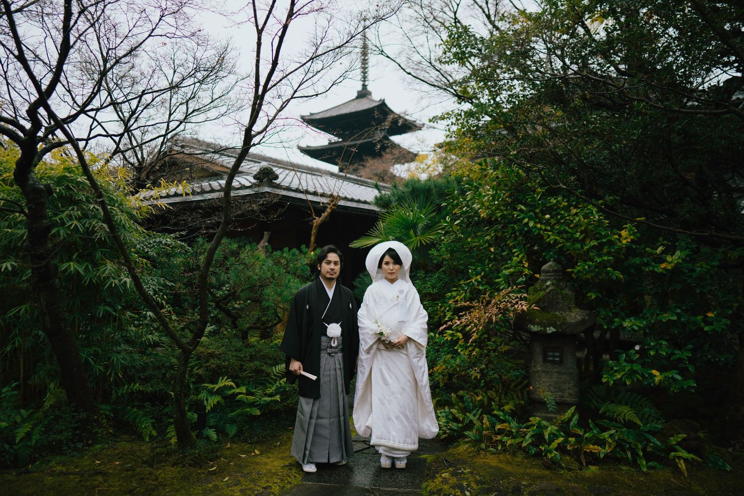 Couple in traditional Japanese wedding attire standing in a lush garden with a pagoda in the background.