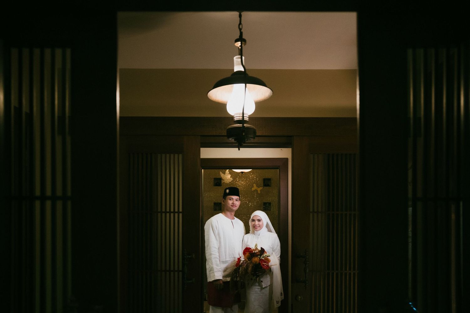 Muslim couple standing in a dimly lit doorway at night, bride holding a red bouquet.