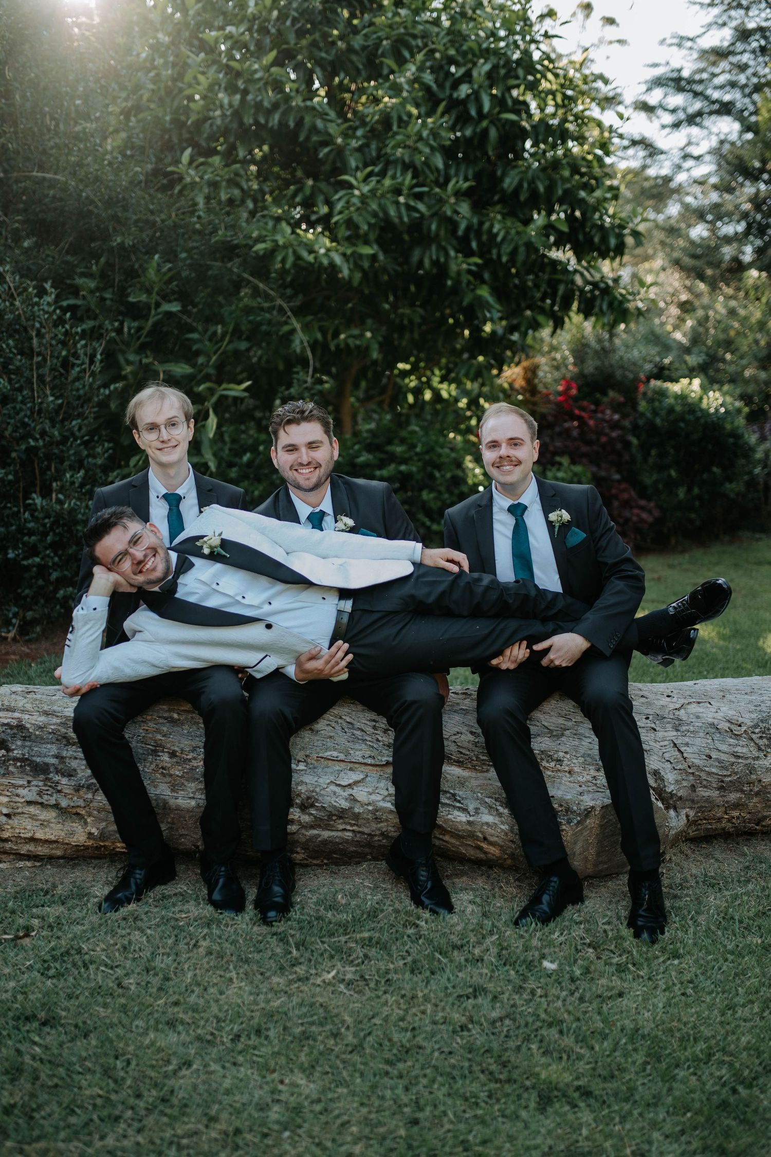 A group in formal attire reclines together on a fallen log in a grassy outdoor setting.