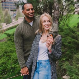 A couple in casual attire standing together outdoors on a green hillside in natural lighting.
