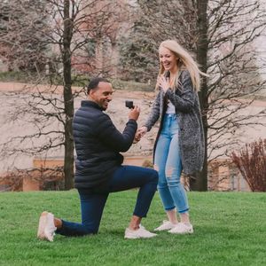 A couple shares a romantic proposal moment on a grassy hillside during a spring day with blooming trees in the background.