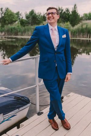 A person in a blue suit and pink tie poses on a dock by a serene lake during sunset.