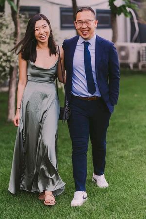 A couple poses together outdoors, with one wearing a sage green dress and the other in a navy suit with white sneakers.