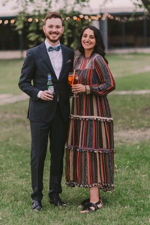 A couple in formal attire shares drinks and smiles while standing together on a grassy lawn at dusk.