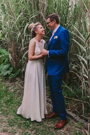 A couple shares a joyful moment in their wedding attire against tall grass, wearing a blue suit and beige gown.