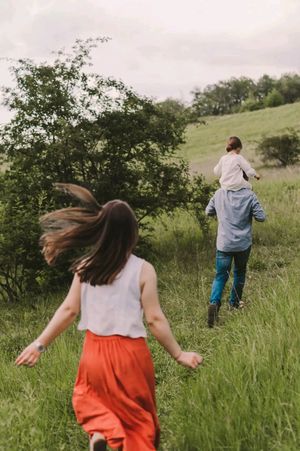 Two people running through a lush green meadow with tall grass and scattered trees on a cloudy summer day.