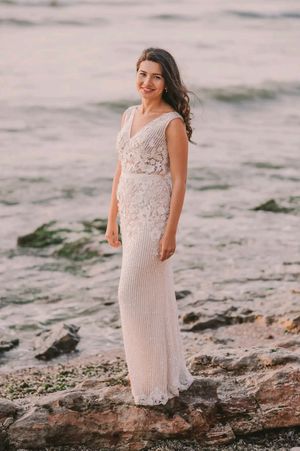 A person in a beaded cream dress poses gracefully on rocky coastal terrain at sunset.