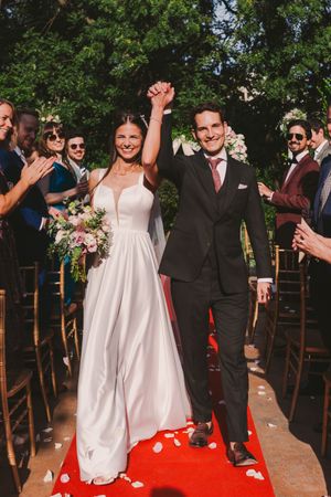 Wedding celebration on a red carpet walkway with guests cheering outdoors.