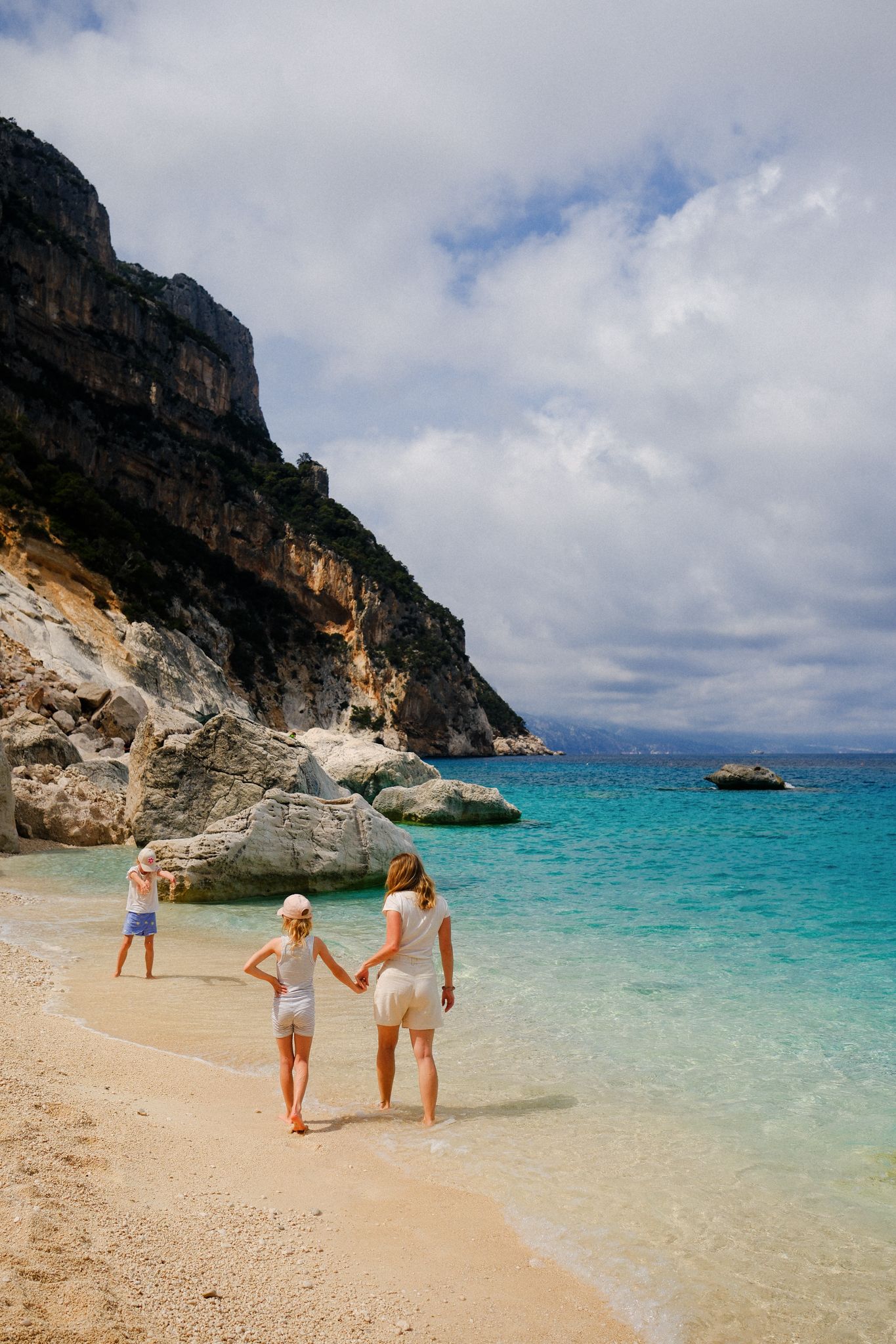 Mother and two children walking hand-in-hand on a stunning turquoise beach with dramatic rocky cliffs in Sardinia.