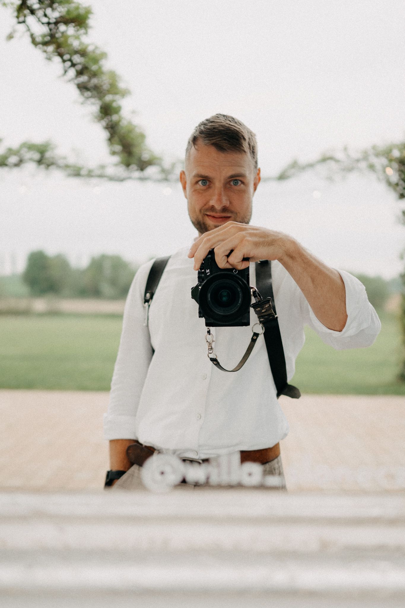 Man in white shirt holding a camera outdoors at a garden event venue with string lights.