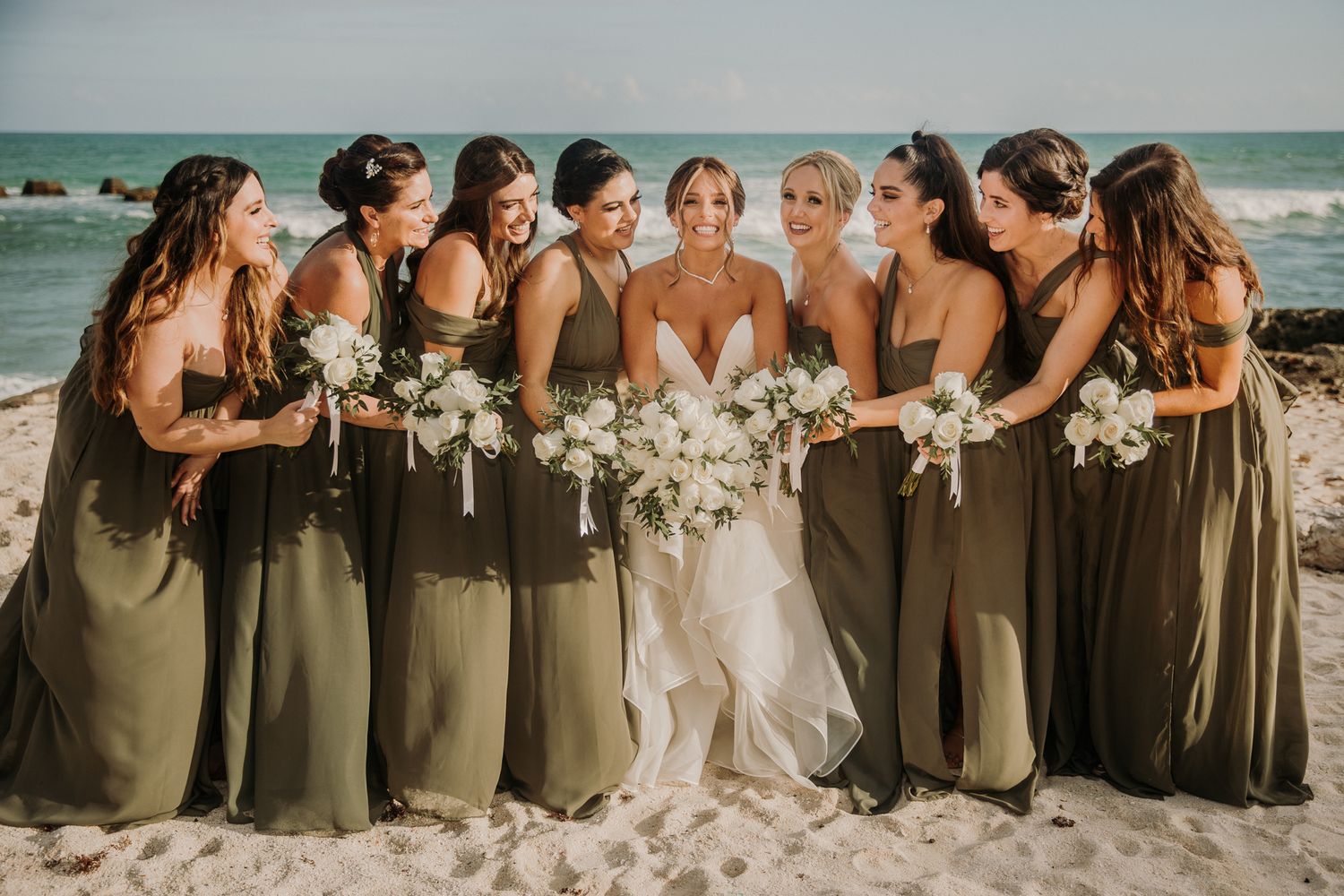 A row of bridesmaids in olive green dresses stand together holding white bouquets on a sunny beach. El Dorado Royale Wedding Photographer
