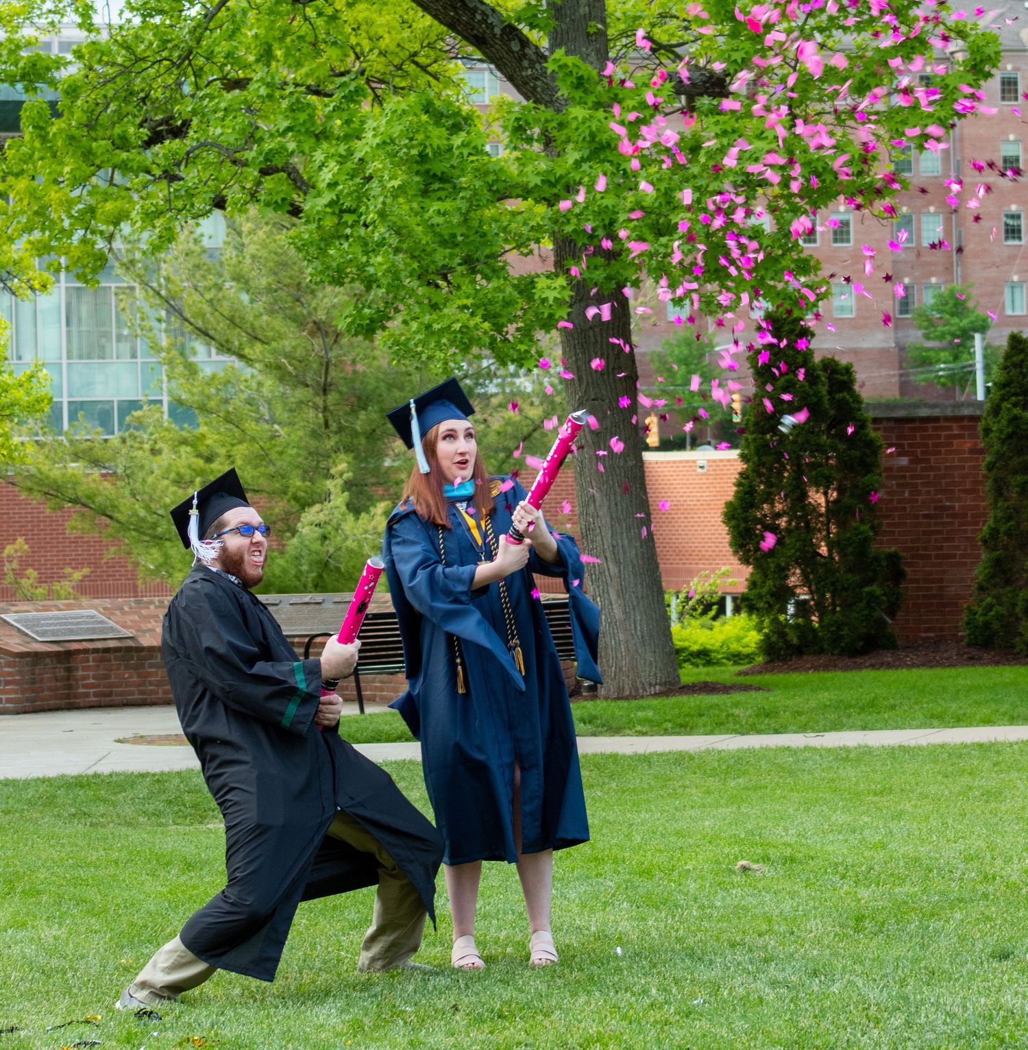 Graduates in black caps and gowns celebrate with playful poses on a sunny spring day with blooming cherry trees.
