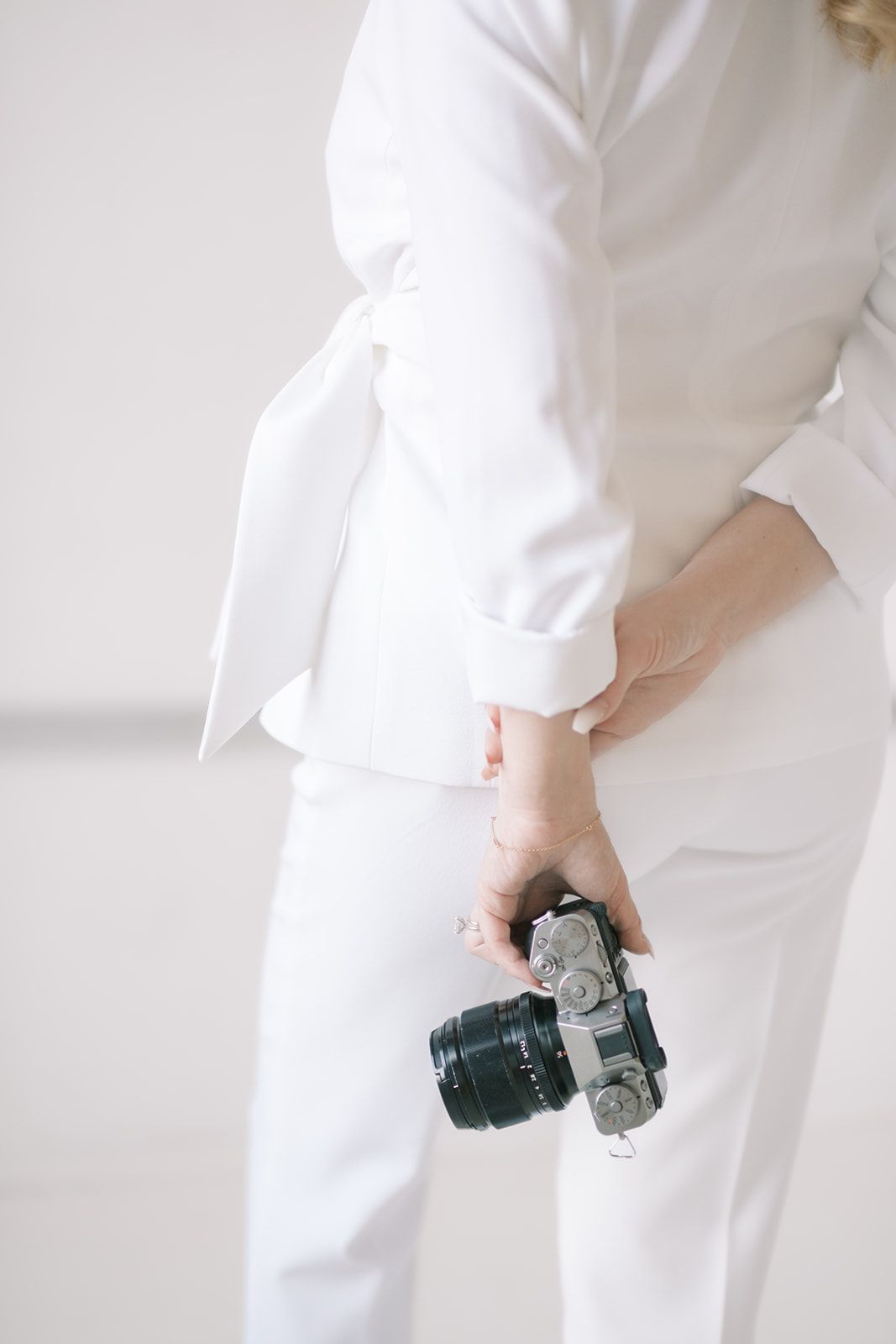 Professional photographer in white outfit holds a DSLR camera against minimal white background.
