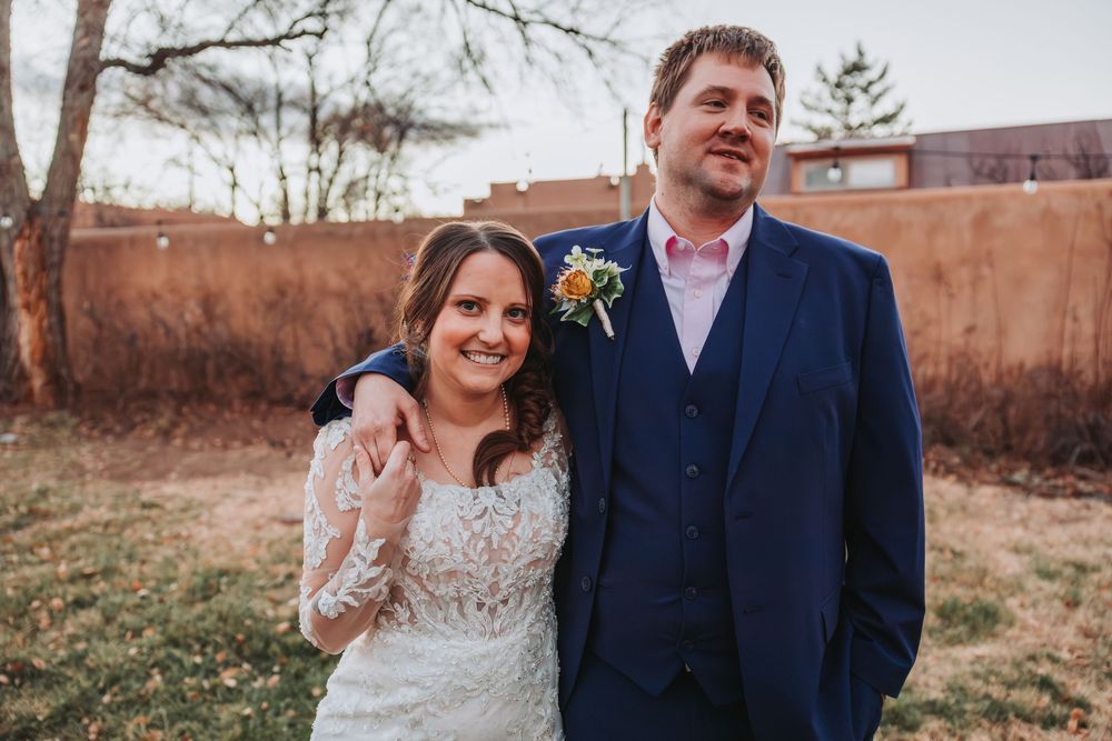 A couple stands together outdoors in wedding attire during a winter day with bare trees in the background.
