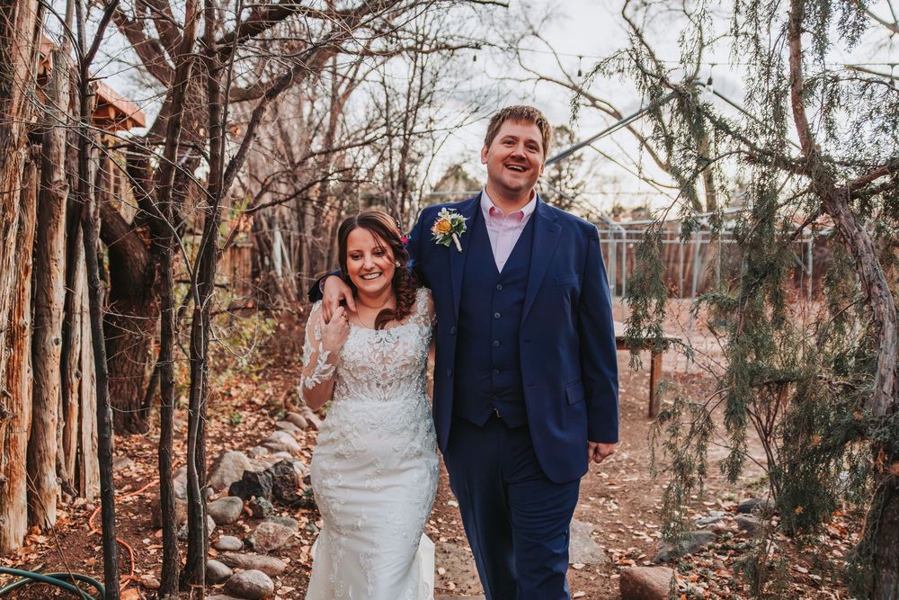 A couple poses together in a wooded area during autumn with trees and fallen leaves surrounding them.
