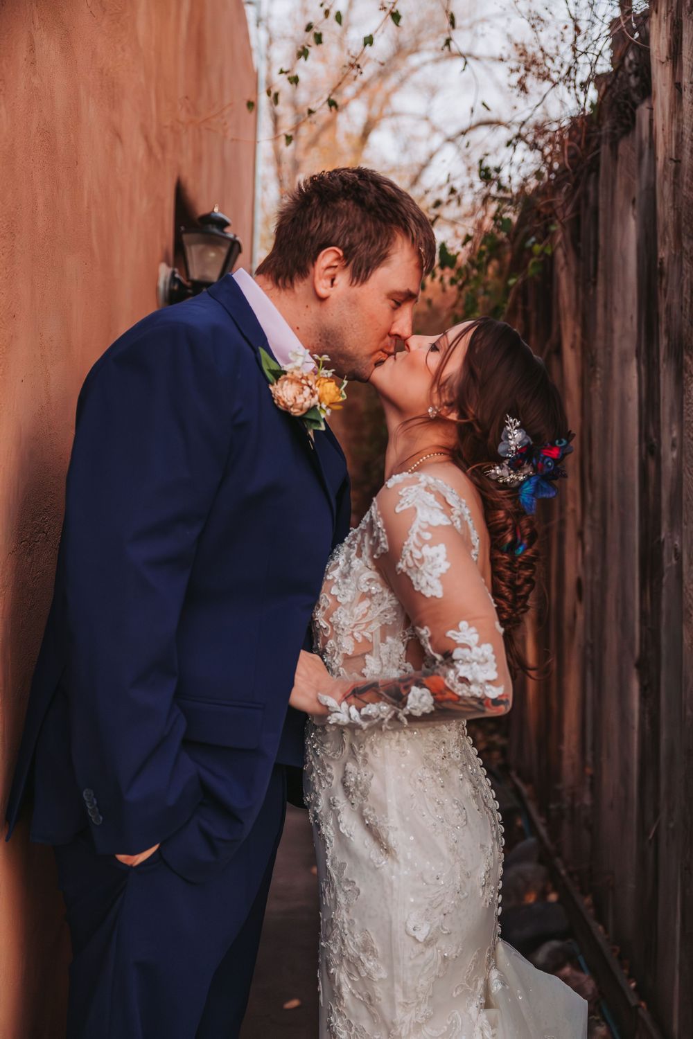 A romantic wedding couple in navy suit and lace dress share intimate kisses against a rustic terracotta wall.