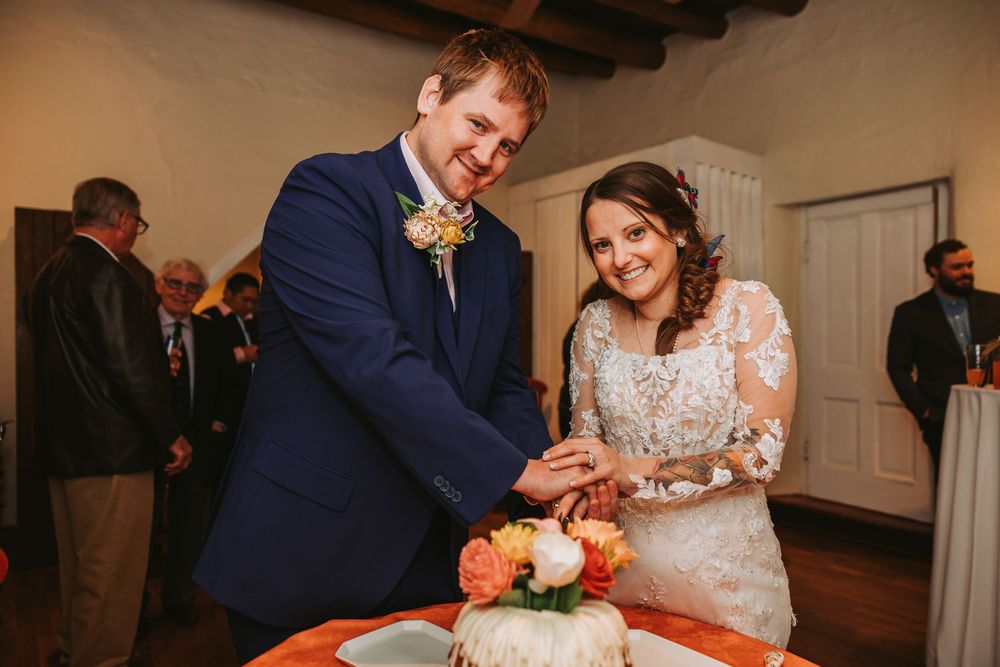 A couple cuts into their orange and white wedding cake together at an indoor reception venue.