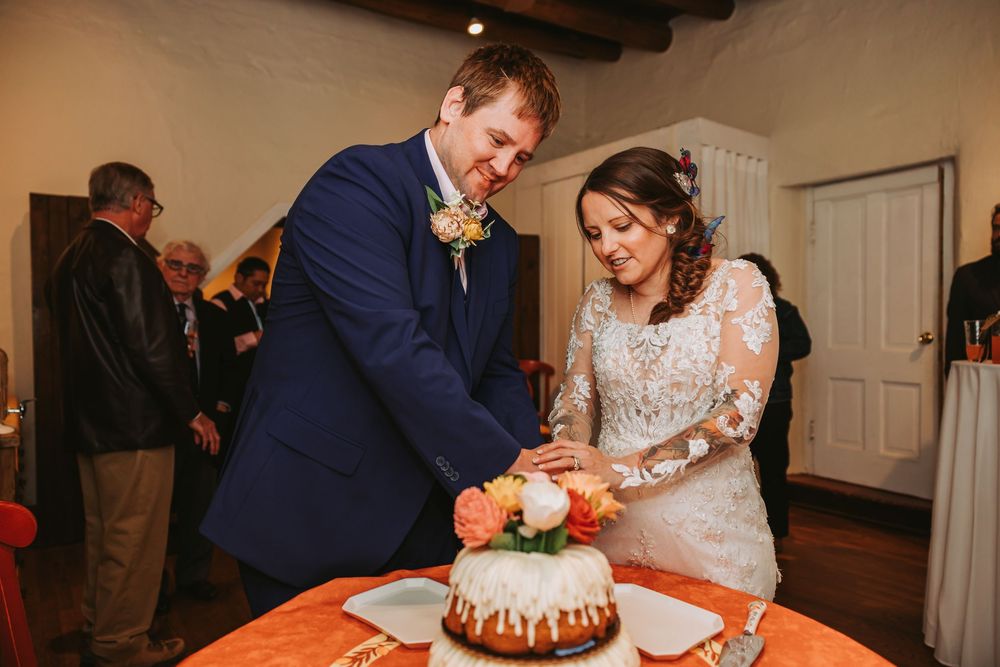 A couple cuts their wedding cake together at their reception while surrounded by guests and floral decorations.