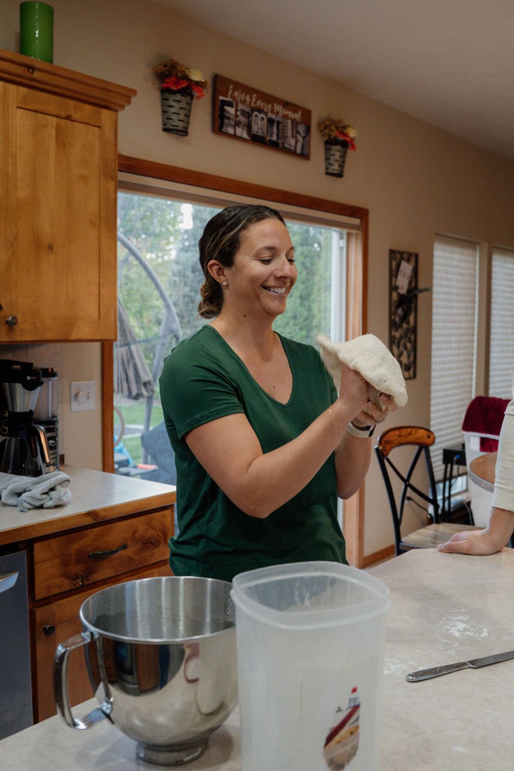 Person in green shirt kneading dough on a floured countertop in a bright kitchen with wooden cabinets.