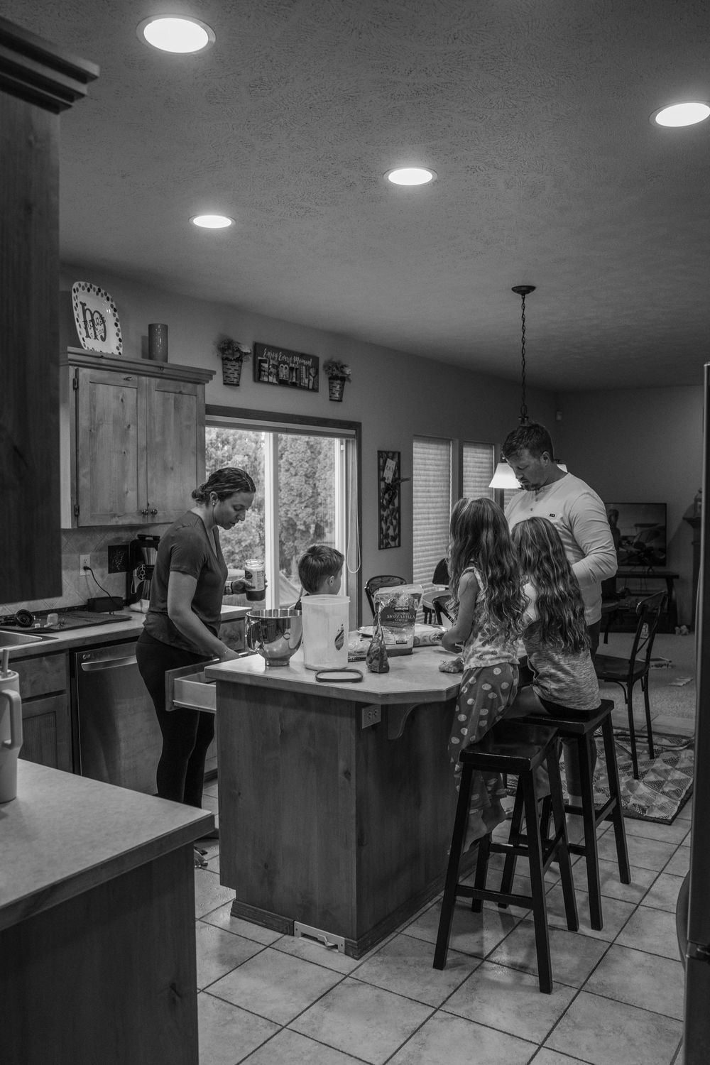 A black and white photo of a family gathering around a kitchen island with recessed lighting overhead.