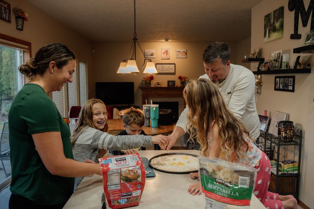 A family gathers around a kitchen table with baking ingredients and pizza boxes.