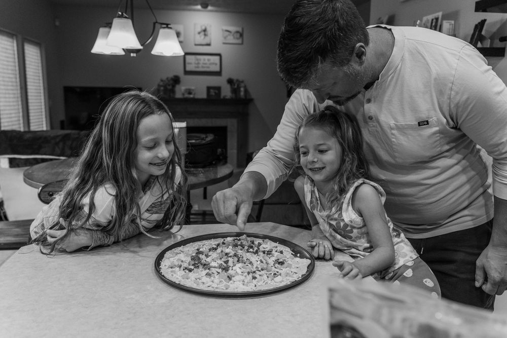 Family members gather around a kitchen counter to prepare and decorate a pizza together.