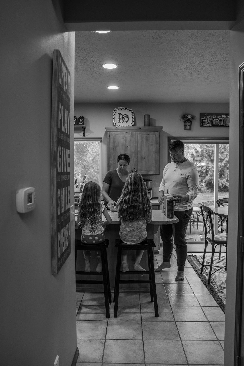 A black and white photograph shows people gathered in a kitchen with tile floors and recessed lighting.