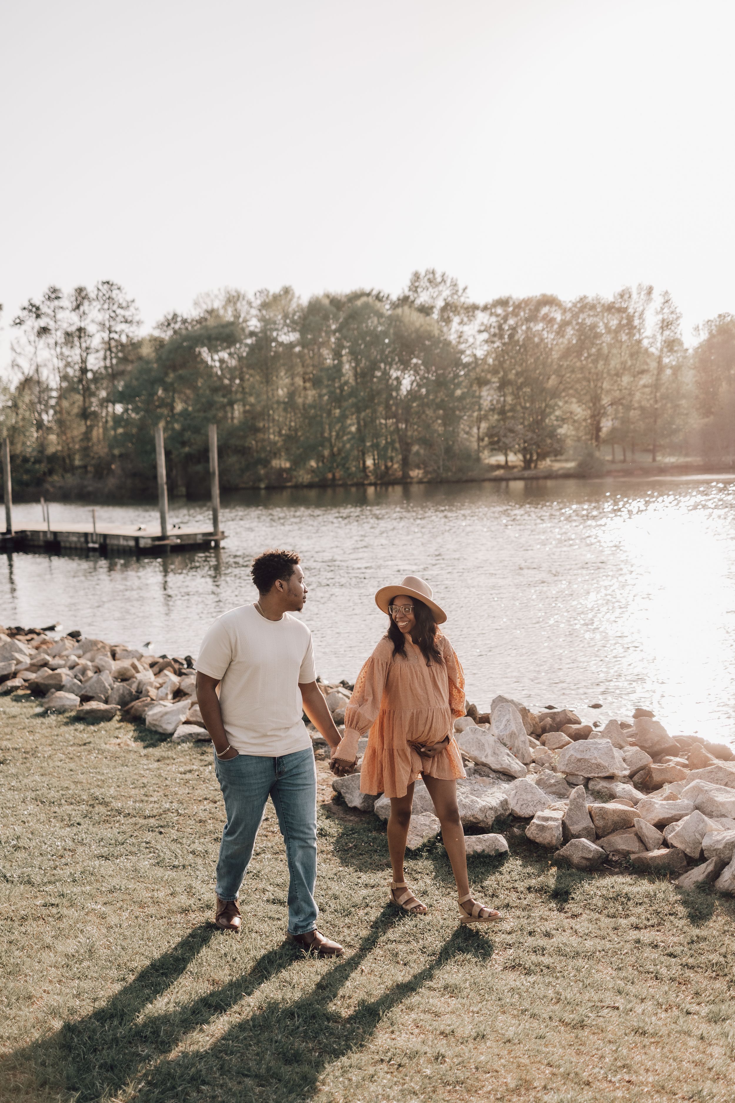 A couple stands by a rocky shoreline at sunset, casting long shadows across the grassy bank.