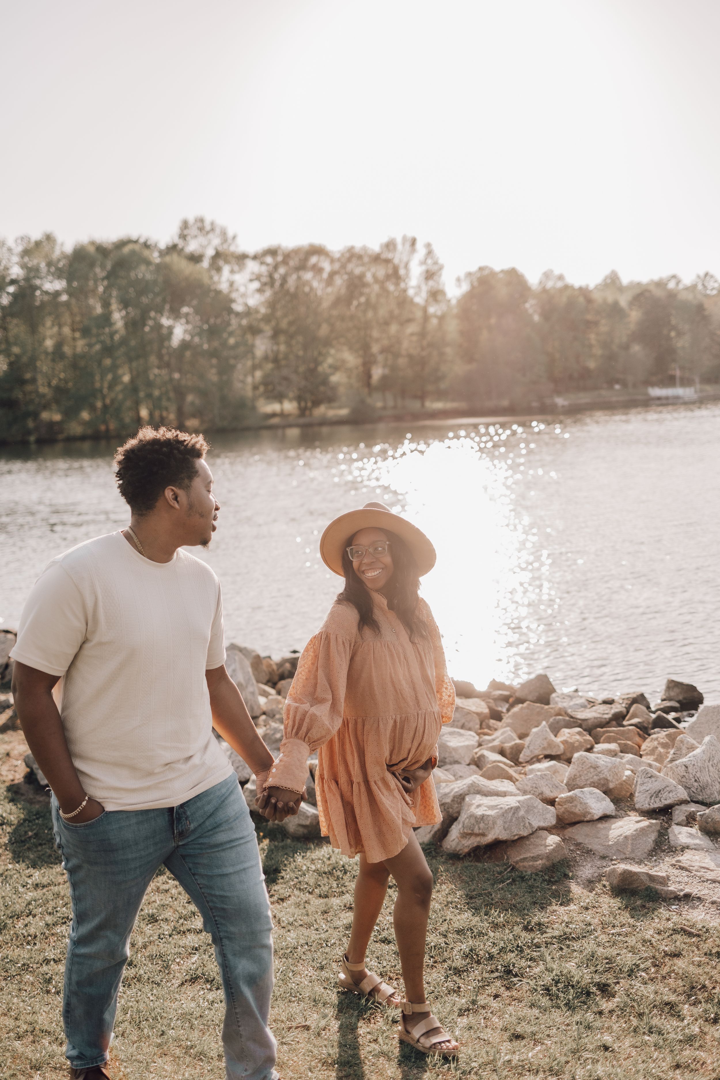 Two people in casual outfits stand together by a lake at sunset, looking out over the water and rocks along the shore.