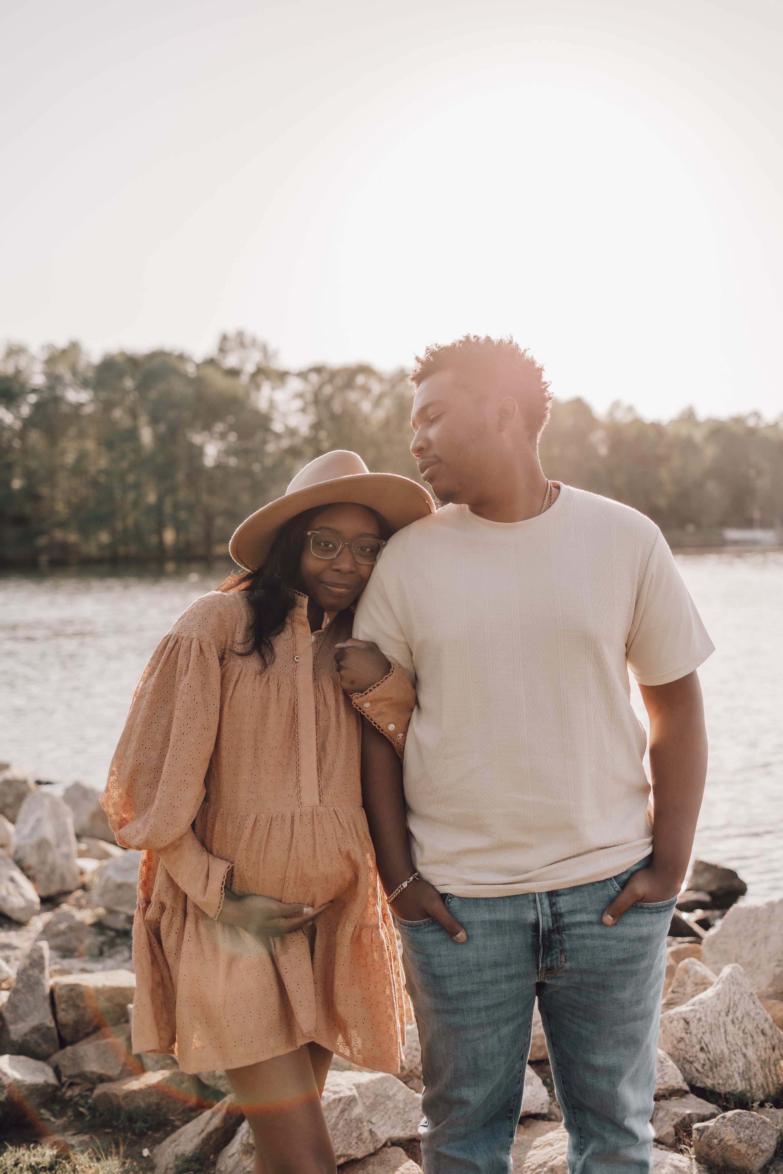 Couple in casual attire sitting together on rocky shoreline at sunset with lake and trees in background.
