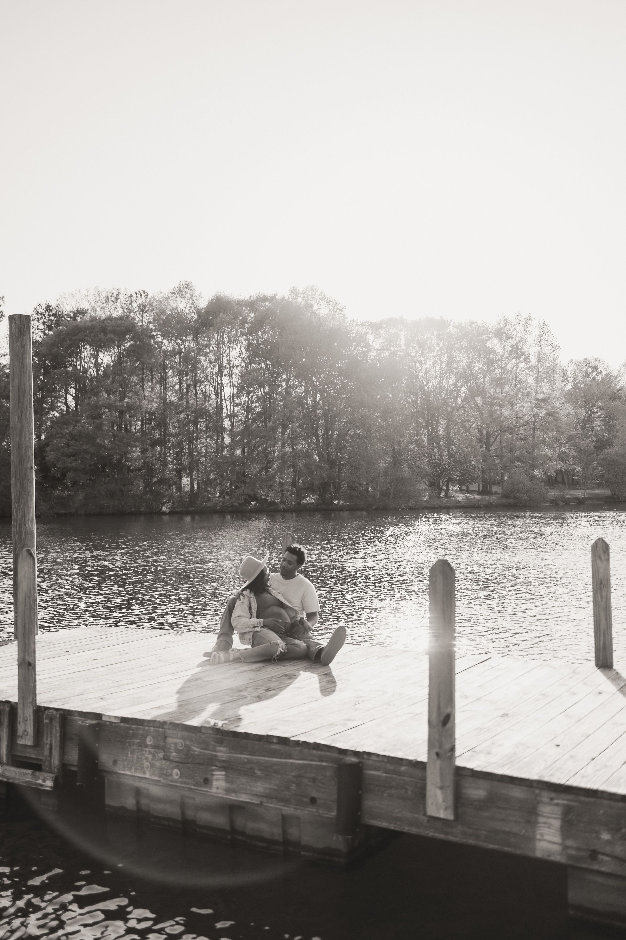 A couple sits together on a wooden dock by a serene lake at sunset, surrounded by silhouetted trees.