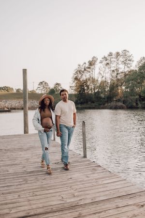 A couple walks together on a wooden dock by a serene lake surrounded by trees at sunset.