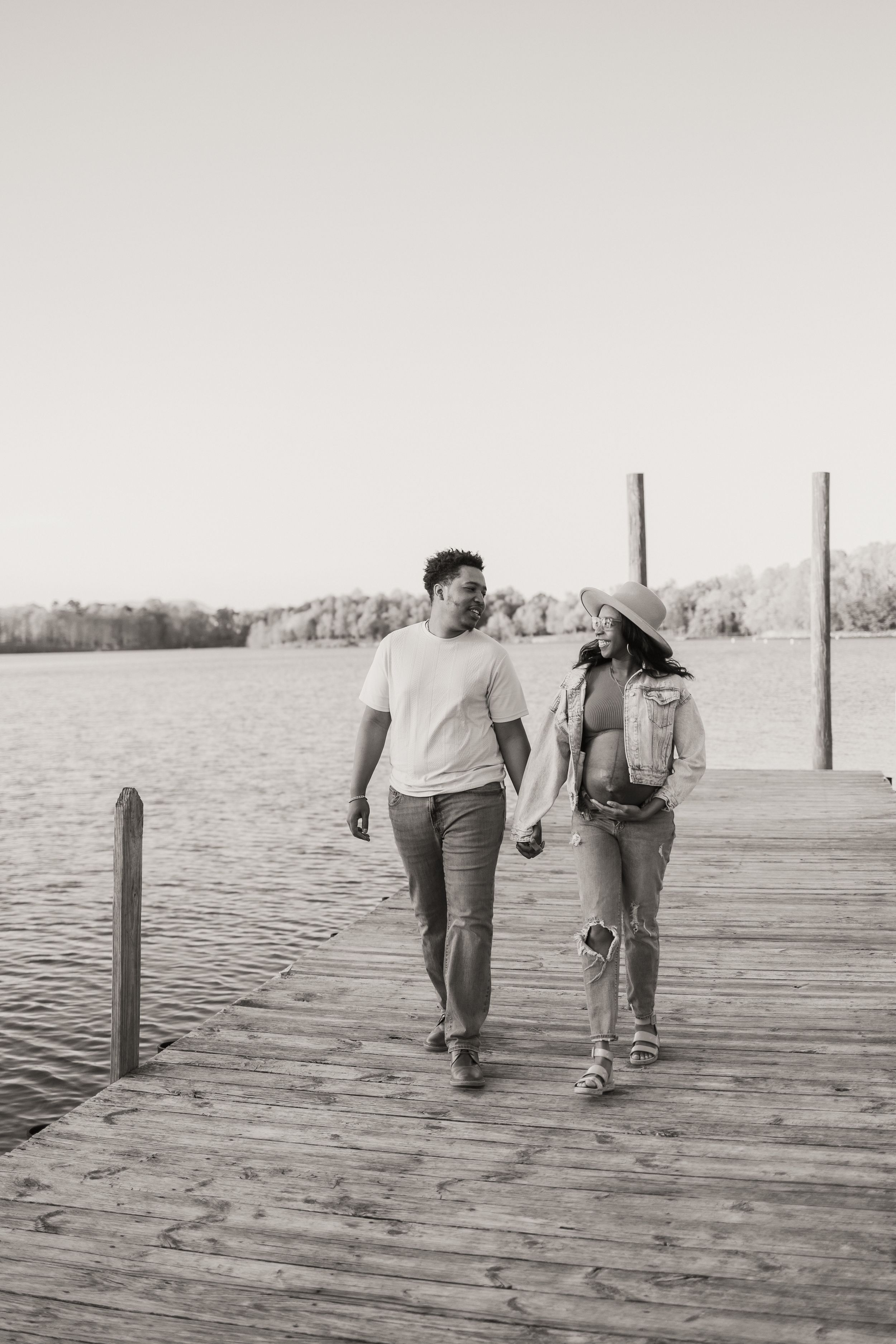A couple walks hand in hand along a wooden dock by the water on a sunny day, captured in black and white.