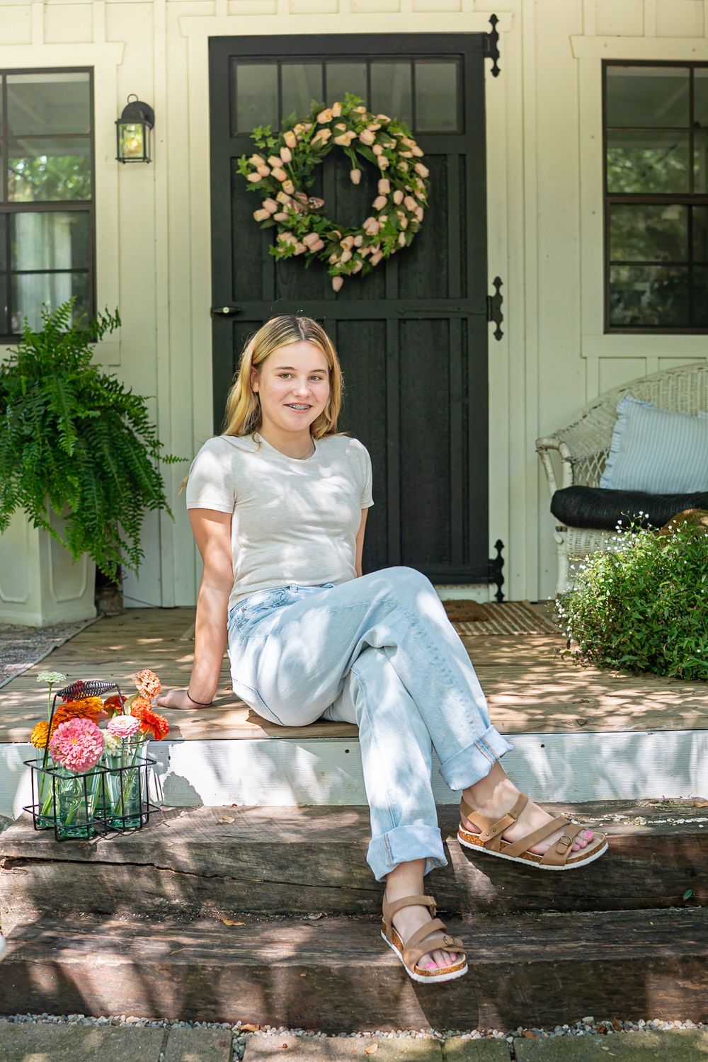 Person sitting on front porch steps with floral wreath and ferns decorating entrance.
