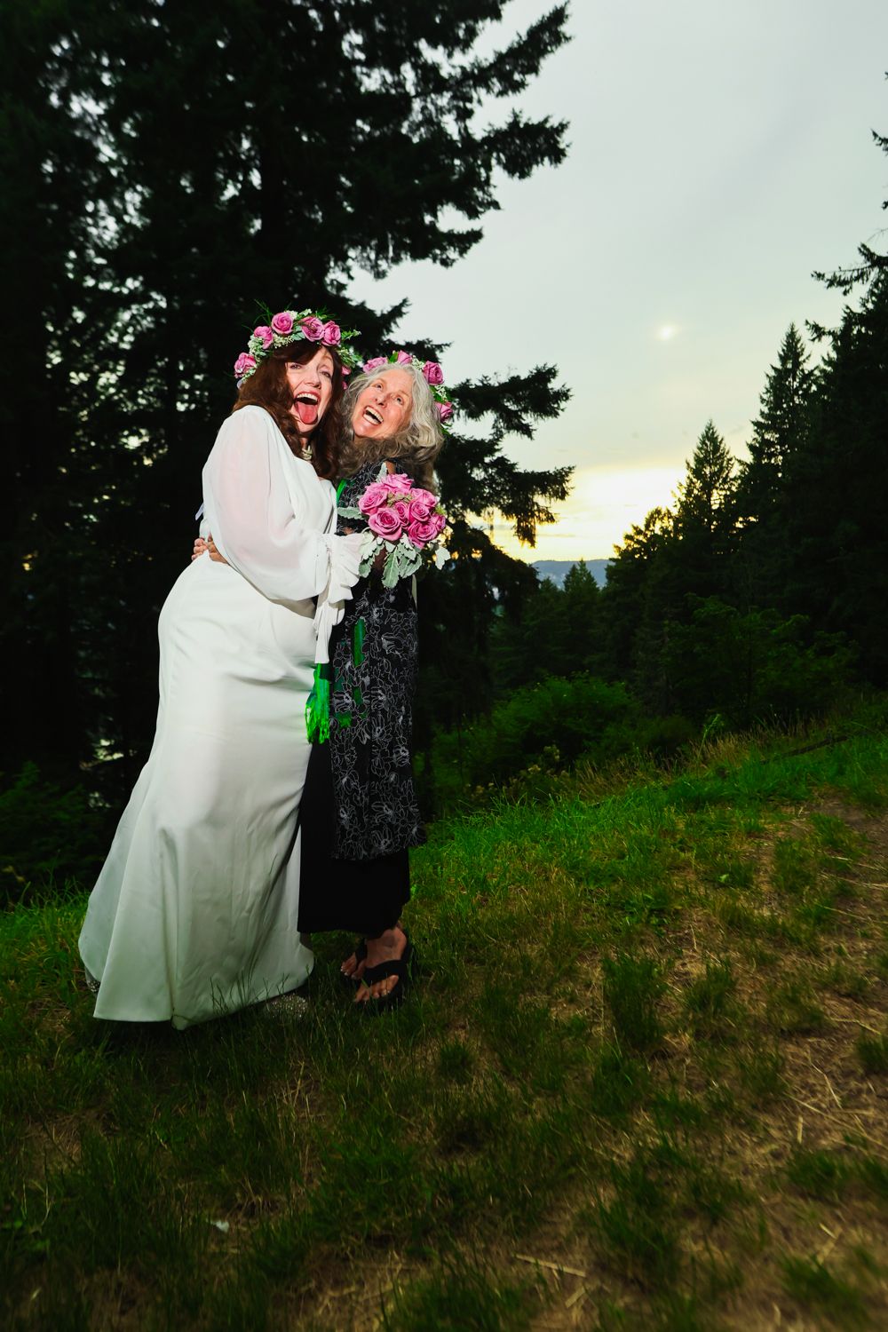 Two people in floral crowns embrace outdoors at sunset with evergreen trees and mountains in the background.