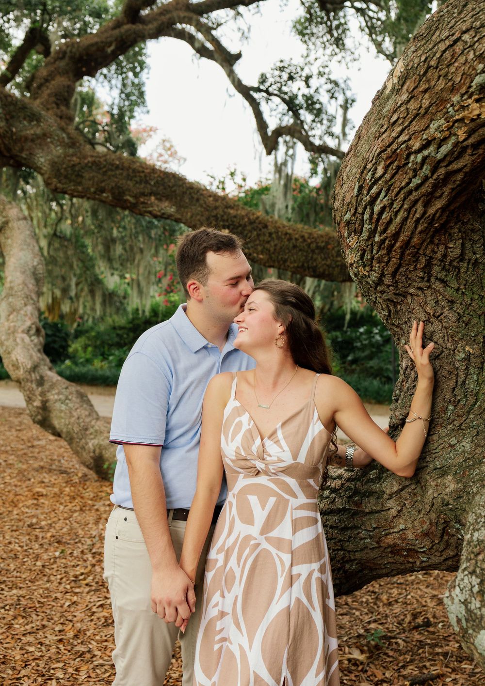 Couple shares a romantic moment by a large oak tree, man kissing woman's forehead as she smiles in a floral dress.