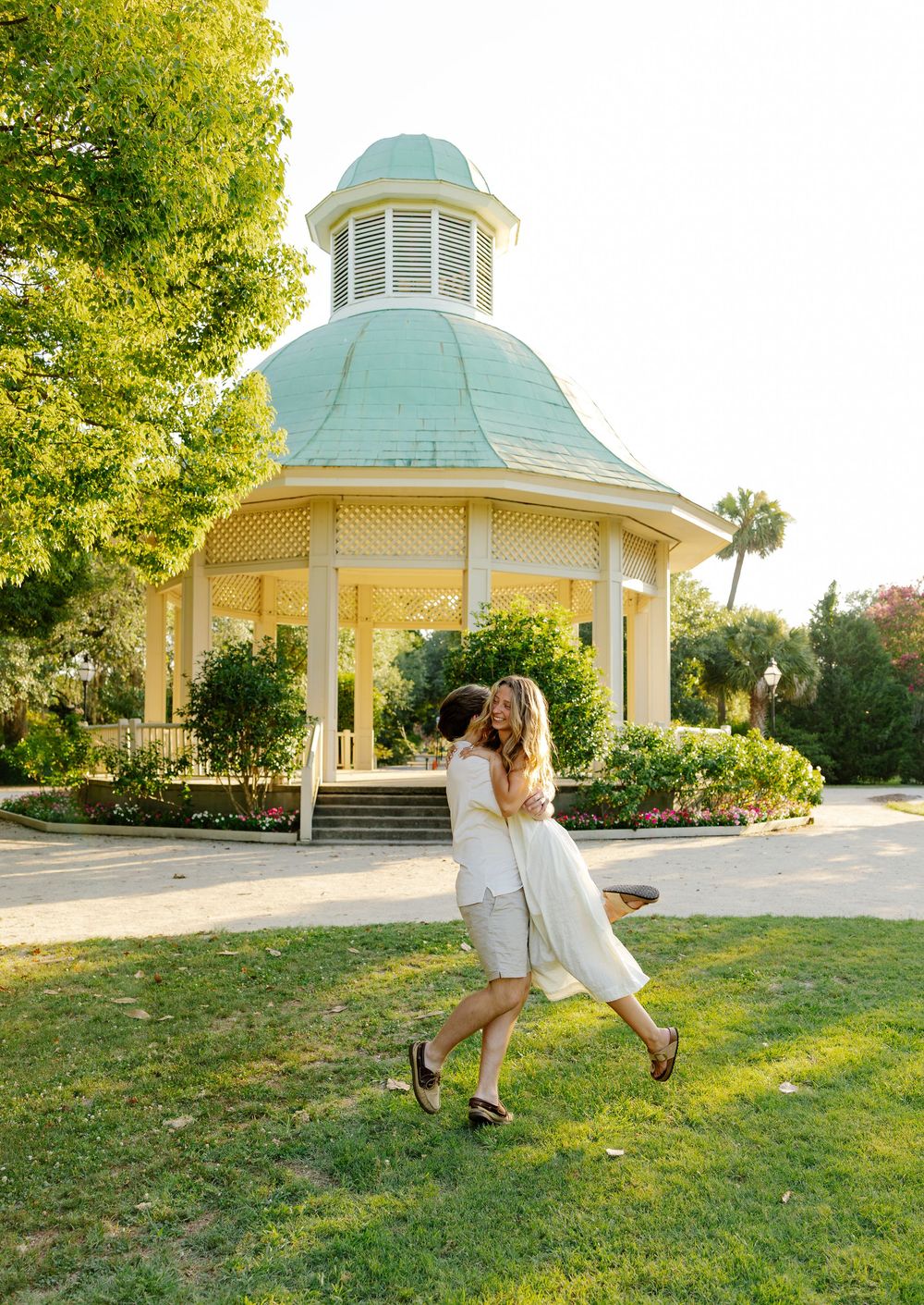 A joyful couple embraces and twirls near a charming yellow gazebo with a green roof in a lush park.