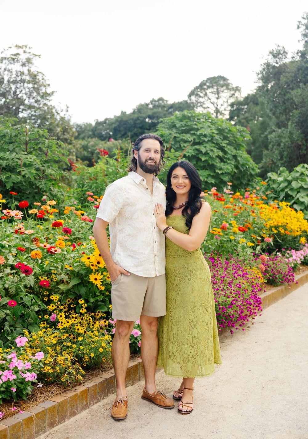 Couple posing together in a lush garden surrounded by colorful flowers, woman in green dress, man in white shirt and khaki shorts.