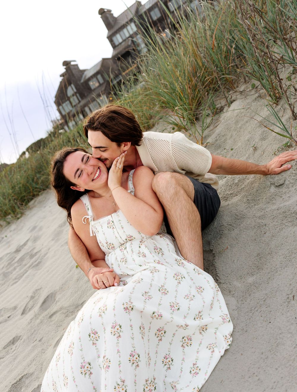 Couple laughing and embracing on a sandy beach dune, woman in floral dress, man kissing her cheek near a coastal home.