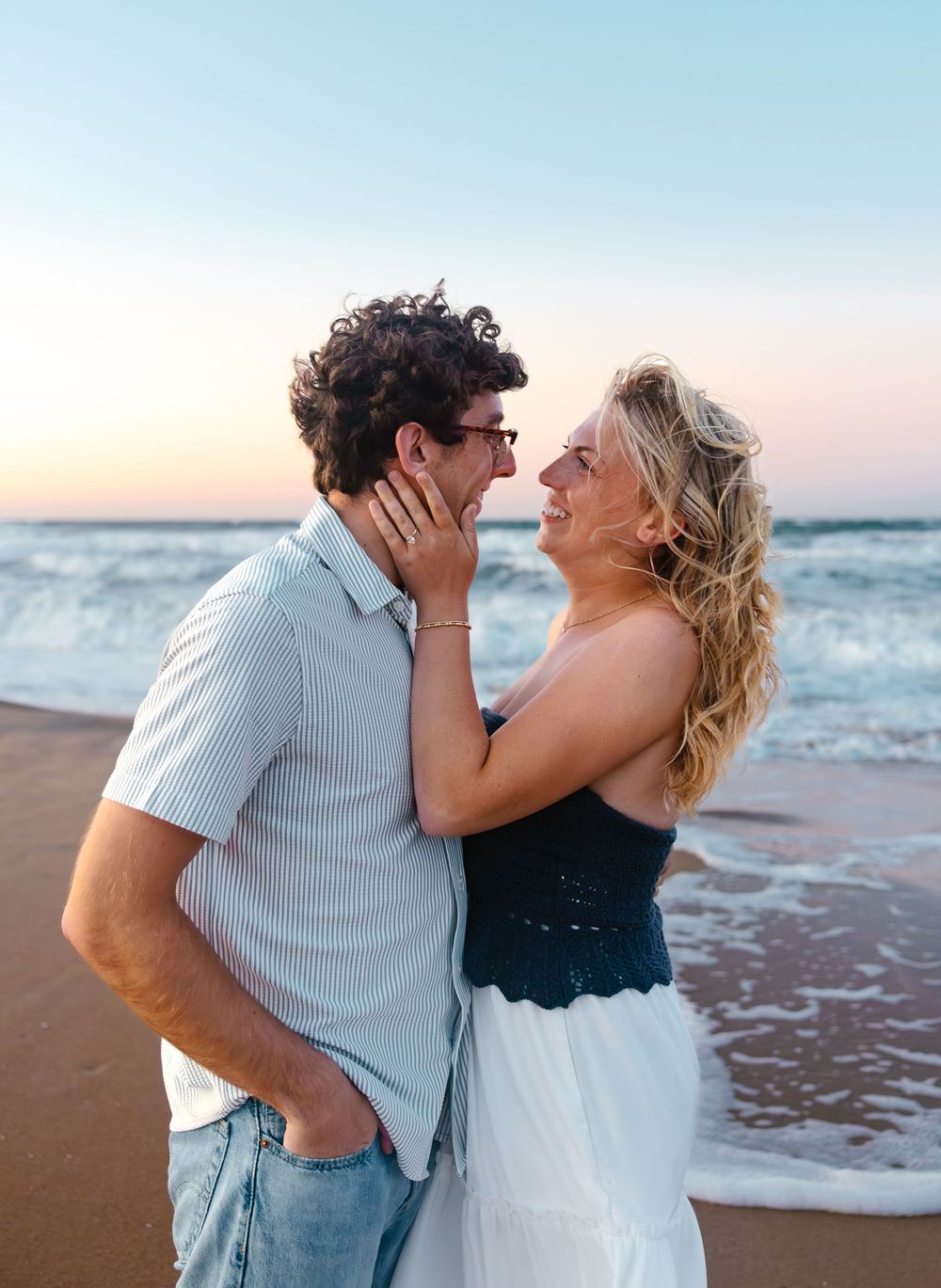 Couple embracing romantically on a sandy beach at sunset, waves crashing behind them, woman in black top and white skirt.