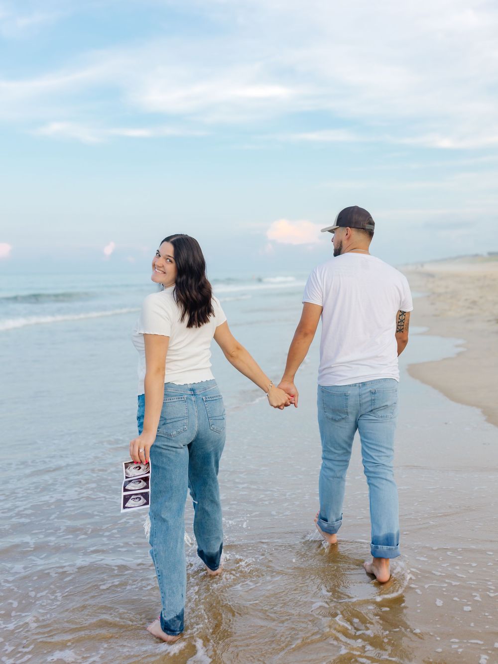 Couple walks hand in hand along beach shoreline in casual denim.