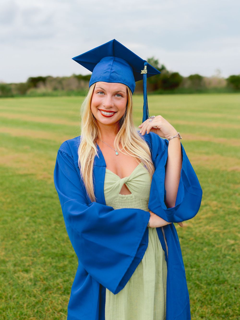 Graduate in blue cap and gown poses in green field during graduation photos.