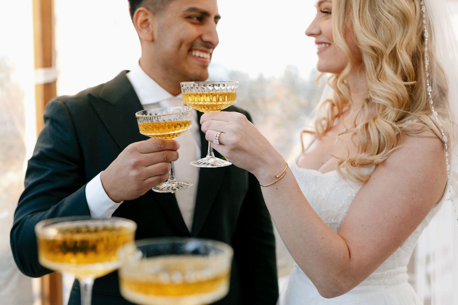 A couple shares a romantic kiss while holding wine glasses at a celebration with string lights in the background.