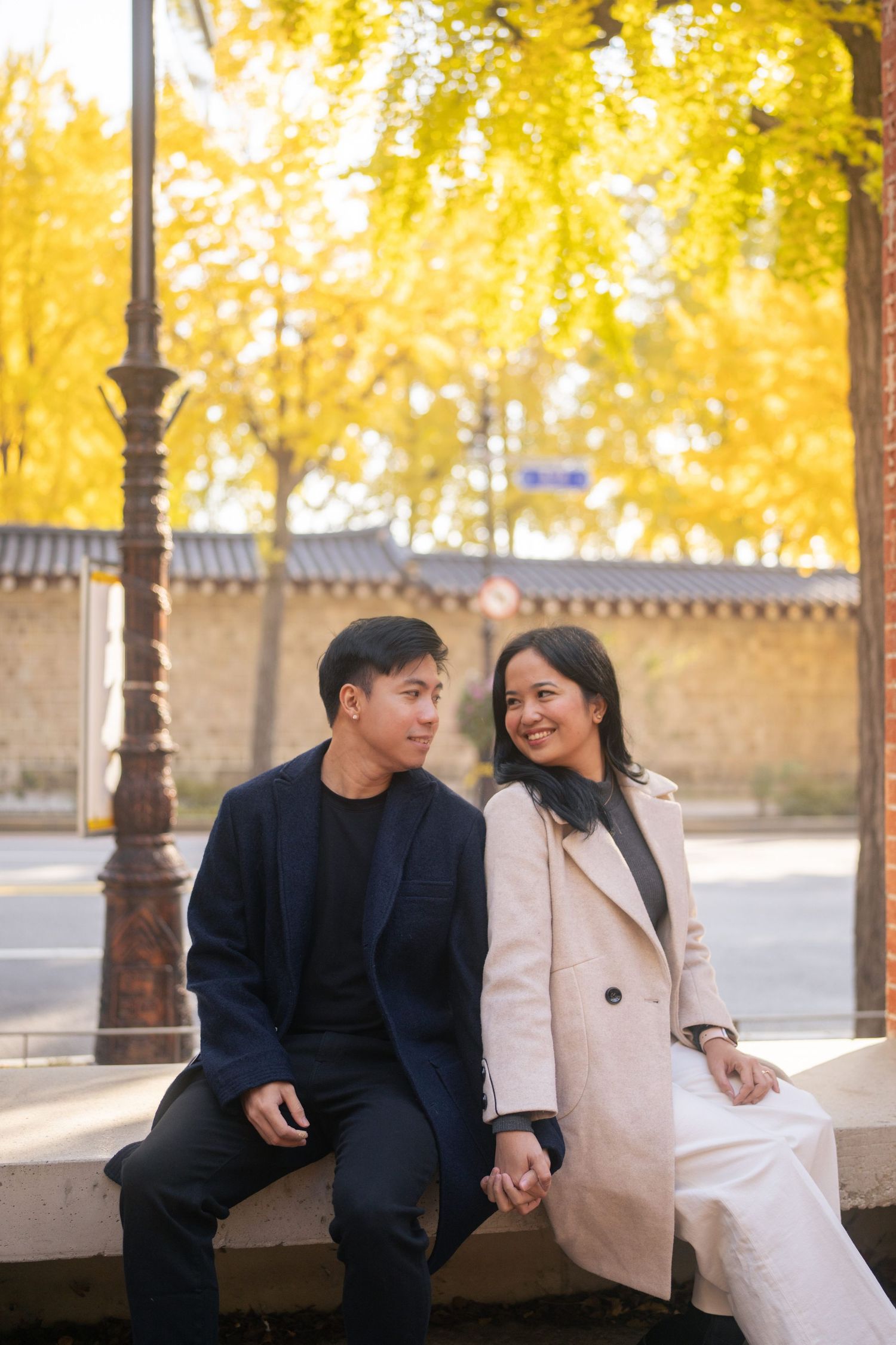 Romantic couple sitting on stone bench under golden autumn trees in urban park setting.