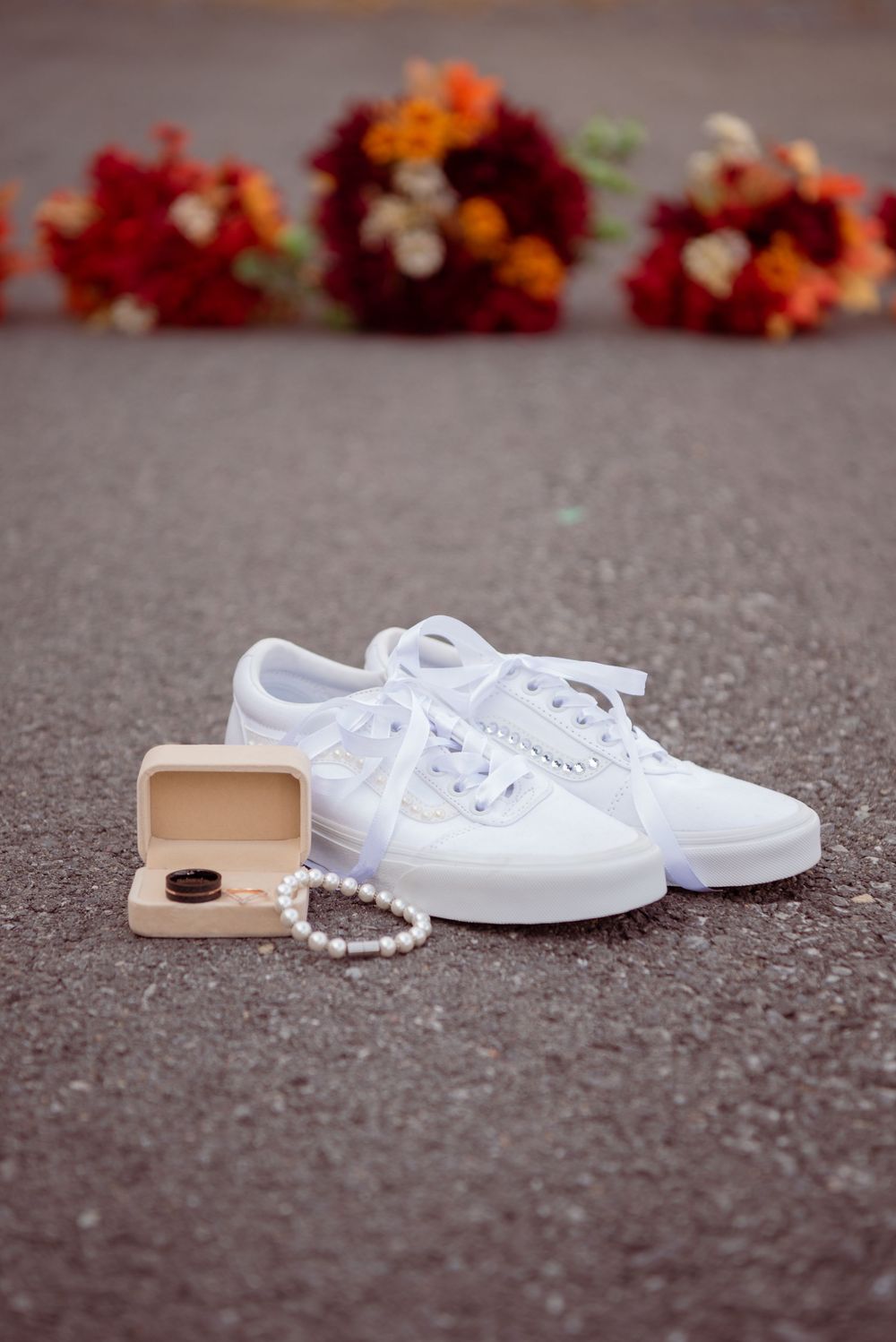 White canvas sneakers with pearls and phone case arranged on asphalt with autumn flowers in background.