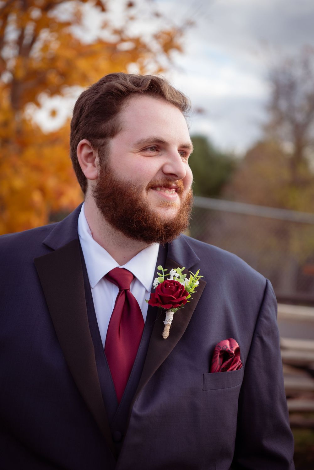 Close-up portrait of groom wearing black suit with burgundy tie and red rose boutonniere against autumn foliage.