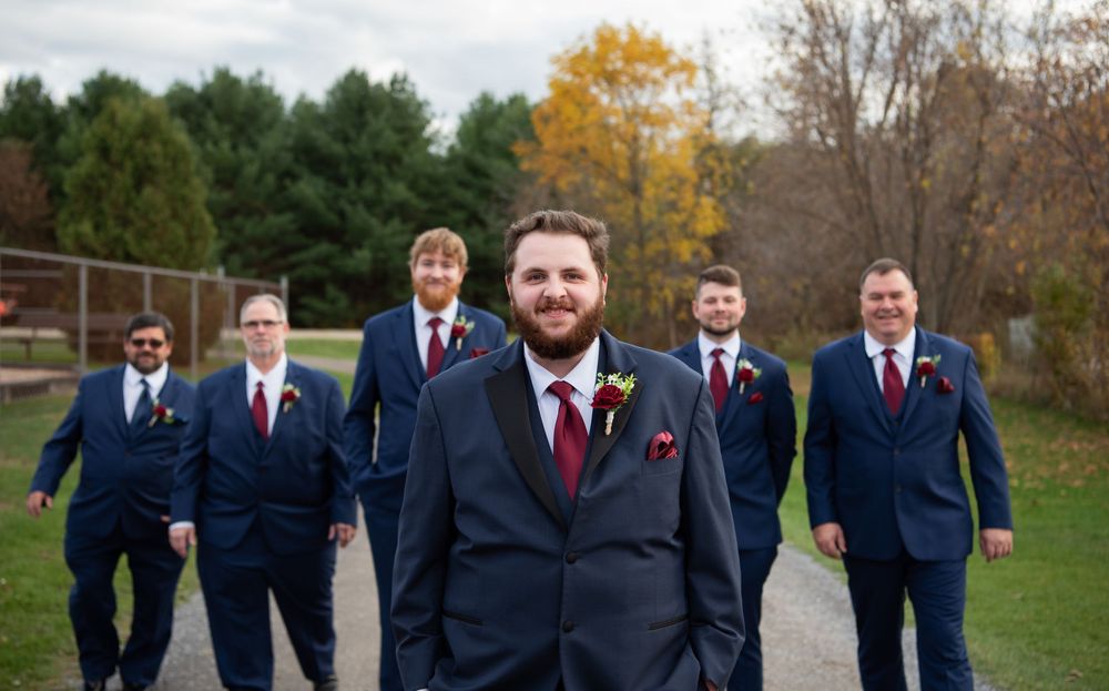 A wedding party in navy suits and burgundy ties poses outdoors during autumn with fall foliage in the background.